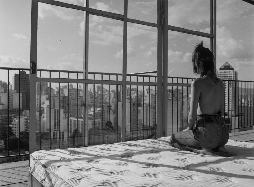 Modelo sentada em pose bucólica olhando para vista da cidade de São Paulo da varanda do apartamento