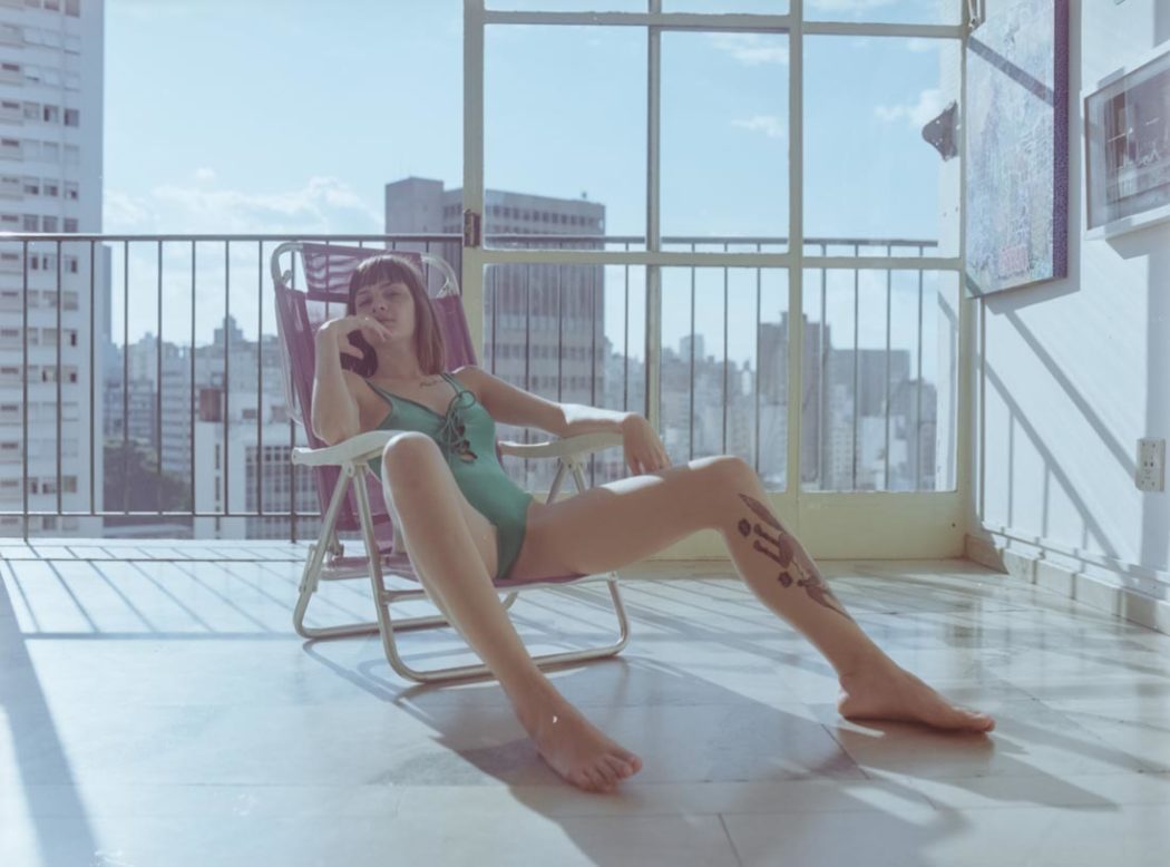 Modelo sentada em cadeira de sol dentro de apartamento amplo com vista para arranha céus