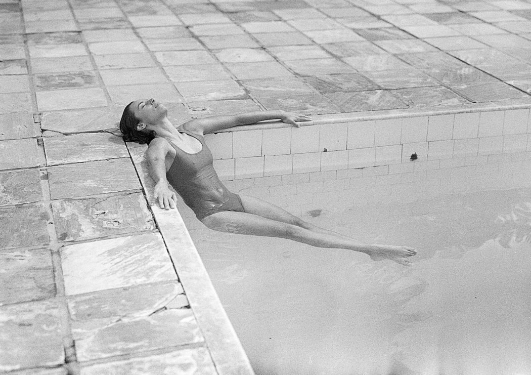 Modelo recostada em borda de piscina com maiô body preto e branco