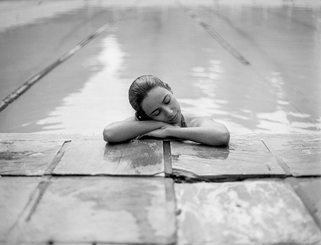 Modelo recostada em borda de piscina com maiô body preto e branco