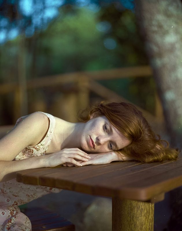 Modelo feminino com expressão bucólica em retrato recostada sobre mesa com cabelo ruivo