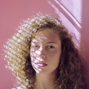 Retrato em close de modelo contra fundo rosa pastel com luz dura de persiana desenhando sombras