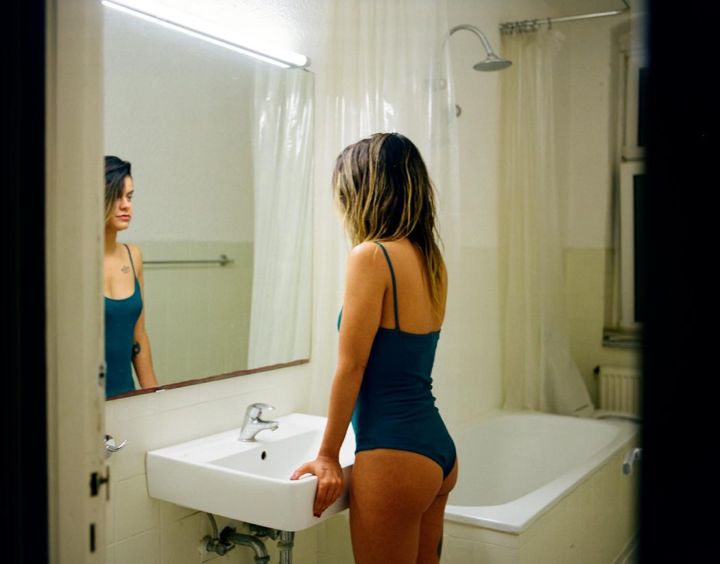 Garota de body maiô diante de espelho em banheiro branco retrô