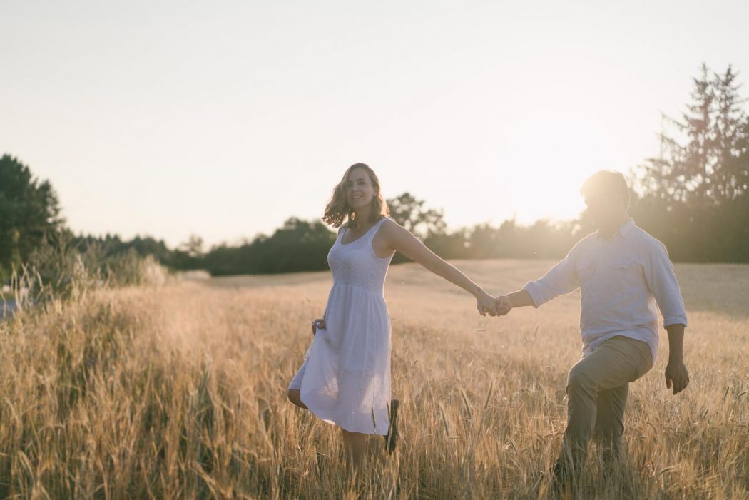 Casal caminhando em campo de trigo amarelo com luz do pôr do sol durante ensaio de casal