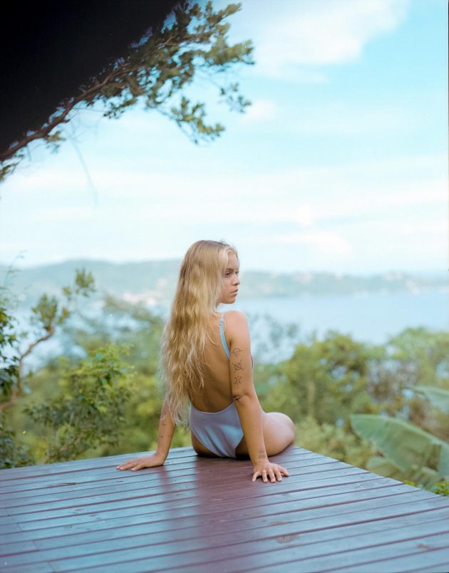 Garota menina loira sentada sobre deck de madeira com vista para lagoa da conceição e céi azul de body maiô