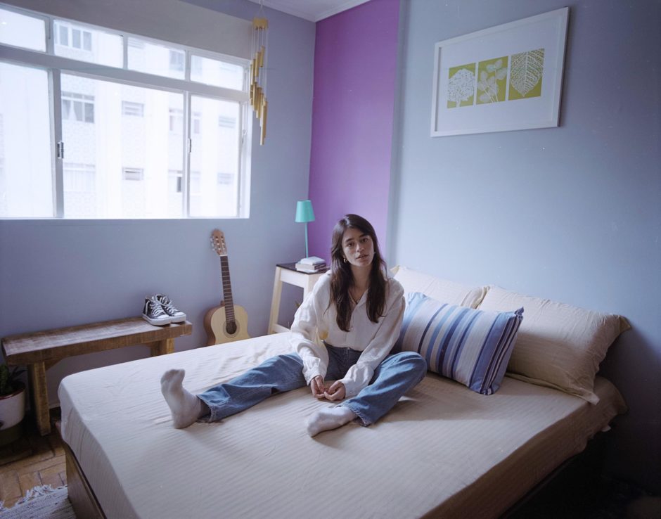 Mulher sentada na cama de pernas abertas com parede em tons azuis e lilás