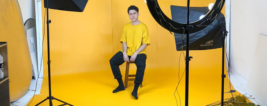 Homem sentando em estúdio fotográfico com fundo amarelo e equipamentos de iluminação