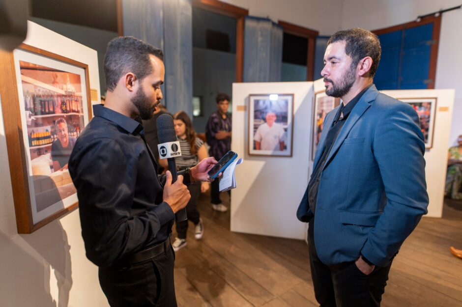 Foto horizontal interna tirada em um ambiente de exposição. À direita, Neto Macedo, vestindo paletó azul-acinzentado sobre camisa preta e calça preta, está em pé com os braços cruzados, olhando para um repórter que o entrevista. O repórter, à esquerda, veste camisa preta e calça preta, e segura um microfone com a marca "INTER TV" em direção a Neto Macedo. O repórter também segura um smartphone e um roteiro. À esquerda deles, em um painel de exposição branco, está uma foto emoldurada de formato vertical mostrando um feirante sorrindo em um mercado de alimentos. Outros painéis com fotos estão visíveis ao fundo, junto com visitantes desfocados e portas rústicas azuis. O chão é de madeira.