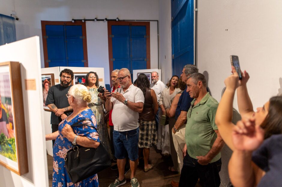 Foto horizontal interna mostrando um corredor de exposição de arte lotado de visitantes. A imagem é tirada por trás de uma pessoa que segura um smartphone no ar para tirar uma foto (primeiro plano à direita). Muitas pessoas estão de pé, aglomeradas e de costas, observando as obras fotográficas expostas em painéis brancos. No primeiro plano à esquerda, uma mulher de vestido azul floral longo e bolsa preta olha para uma foto emoldurada de um feirante. Vários outros visitantes, de diferentes idades e vestindo roupas casuais, preenchem o espaço, olhando para as fotos ou para seus próprios celulares. O ambiente do museu apresenta portas rústicas azuis ao fundo e chão de madeira.