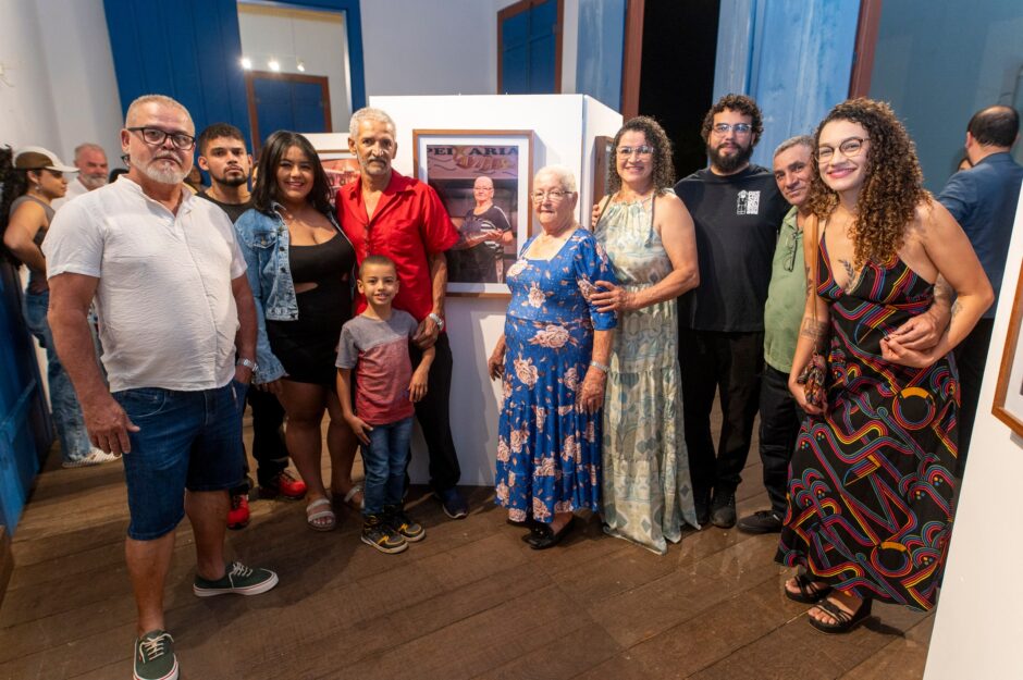 Foto horizontal interna de um grande grupo de pessoas posando para uma foto de família em frente a um retrato emoldurado na exposição. Dez pessoas, de várias idades, estão reunidas em torno de um painel de exposição branco que exibe uma foto vertical de uma feirante idosa. No centro do grupo, a feirante idosa na vida real (vestindo um vestido longo azul floral) está em pé, tocando sua própria obra. Seus familiares e amigos a cercam. Da esquerda para a direita: um homem de camisa polo branca e bermuda jeans; um jovem; uma mulher de jaqueta jeans; um homem de camisa vermelha; um menino pequeno; a feirante idosa; uma mulher de vestido longo estampado; um homem de barba e camiseta preta; um homem de camisa polo verde; e uma mulher de vestido longo estampado colorido e óculos. Todos sorriem para a câmera. O ambiente da exposição com portas rústicas azuis e chão de madeira é visível ao fundo.
