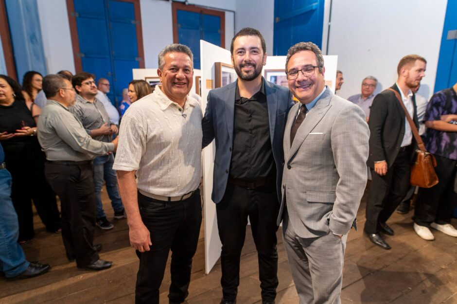 Uma foto de plano médio tirada em uma recepção de exposição de arte. No centro, três homens estão em pé, sorrindo para a câmera. Da esquerda para a direita: Valmir, um feirante retratado, vestindo camisa bege e calças escuras; Neto Macedo, o fotógrafo, vestindo paletó azul-acinzentado sobre camisa preta; e Wagner de Paulo Santiago, reitor da Unimontes, vestindo um terno cinza texturizado e gravata. Neto Macedo tem os braços em volta dos ombros dos outros dois homens. O fundo mostra painéis de exposição brancos com fotos emolduradas, portas rústicas azuis e outros convidados desfocados.